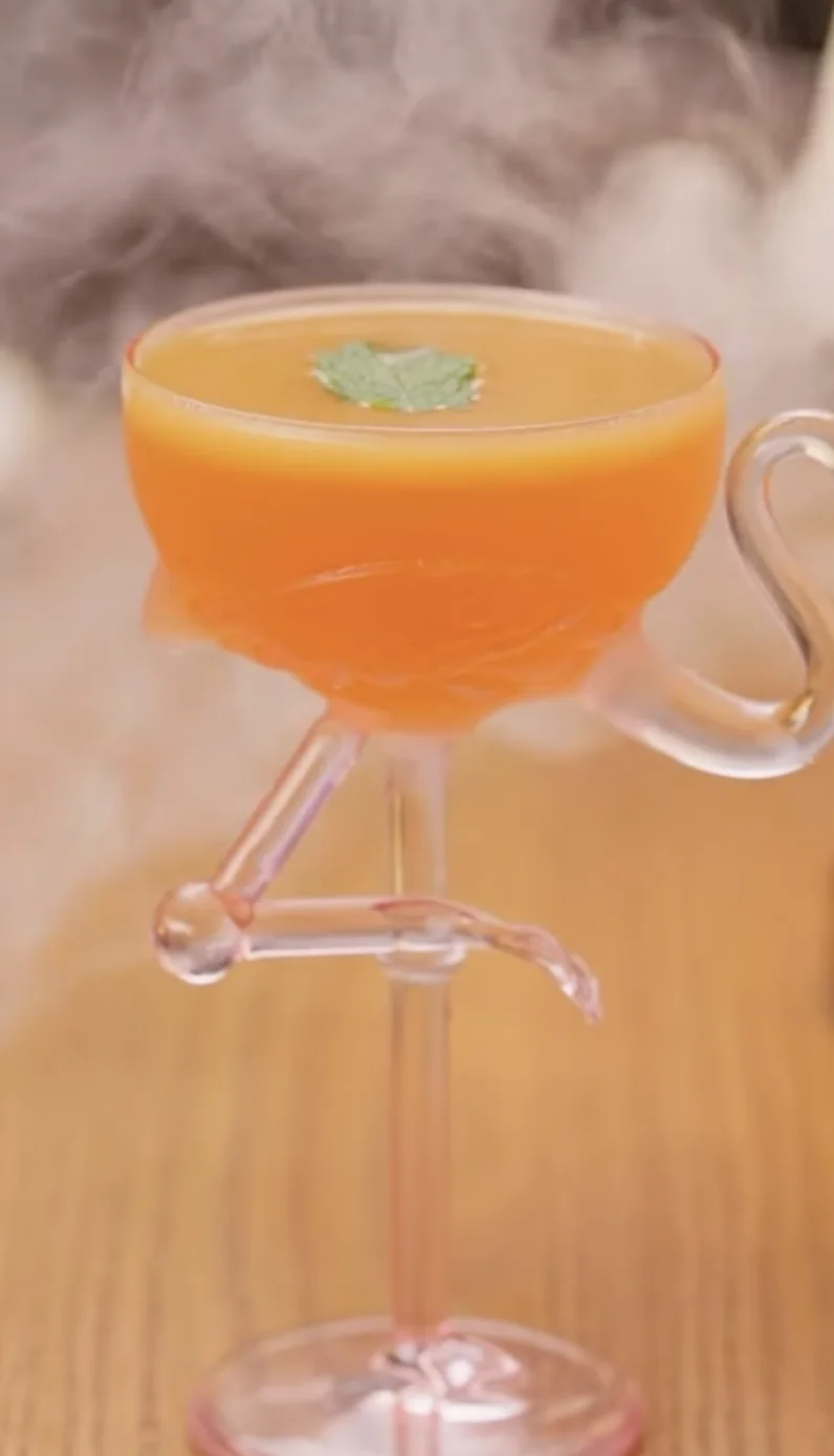 An orange cocktail in a flamingo coupe surrounded by mist
